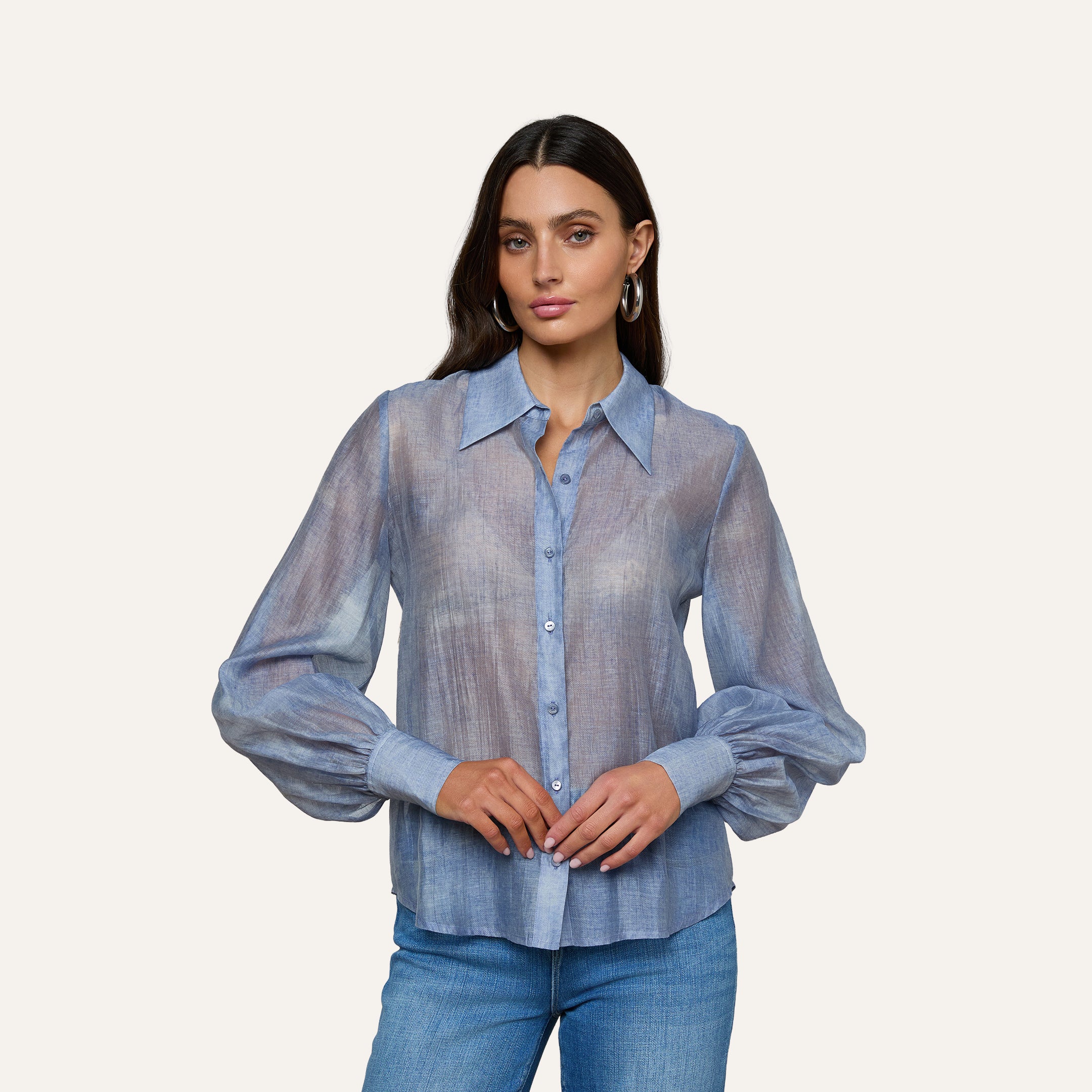 A woman with long dark hair wears the Jayleen Blouse—a light blue sheer chiffon top with blouson sleeves—paired with blue jeans, standing against a plain light gray background.