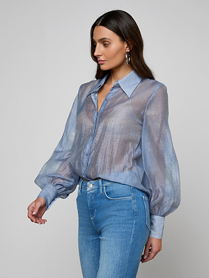 A woman with long dark hair wears the Jayleen Blouse, a sheer chiffon button-down with blouson sleeves, paired with light blue jeans. She stands against a plain gray background, looking to the side.