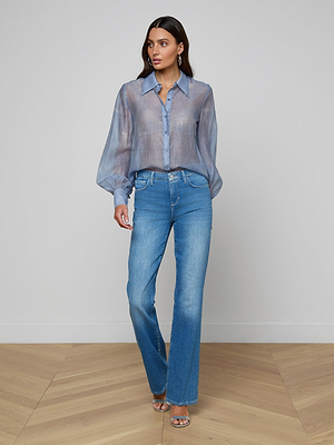 A woman stands indoors on a wooden floor, wearing the Jayleen Blouse with blouson sleeves, paired with blue jeans and silver high-heeled sandals. Her hair is down and her hands are in her pockets.