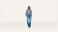 A woman stands against a plain white background, wearing a sheer blue long-sleeve shirt, blue jeans, and open-toed heels.