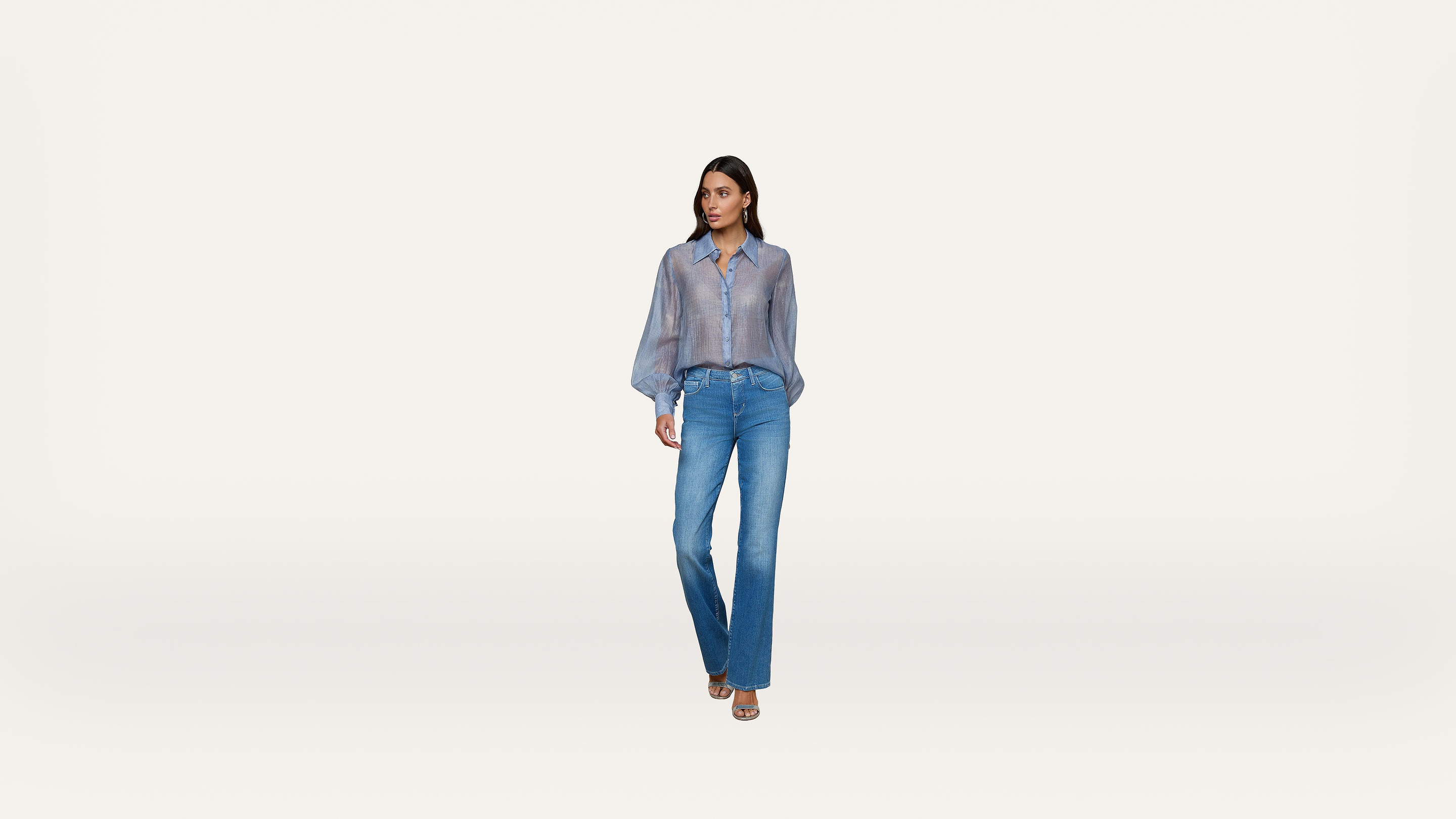 A woman stands against a plain white background, wearing a sheer blue long-sleeve shirt, blue jeans, and open-toed heels.