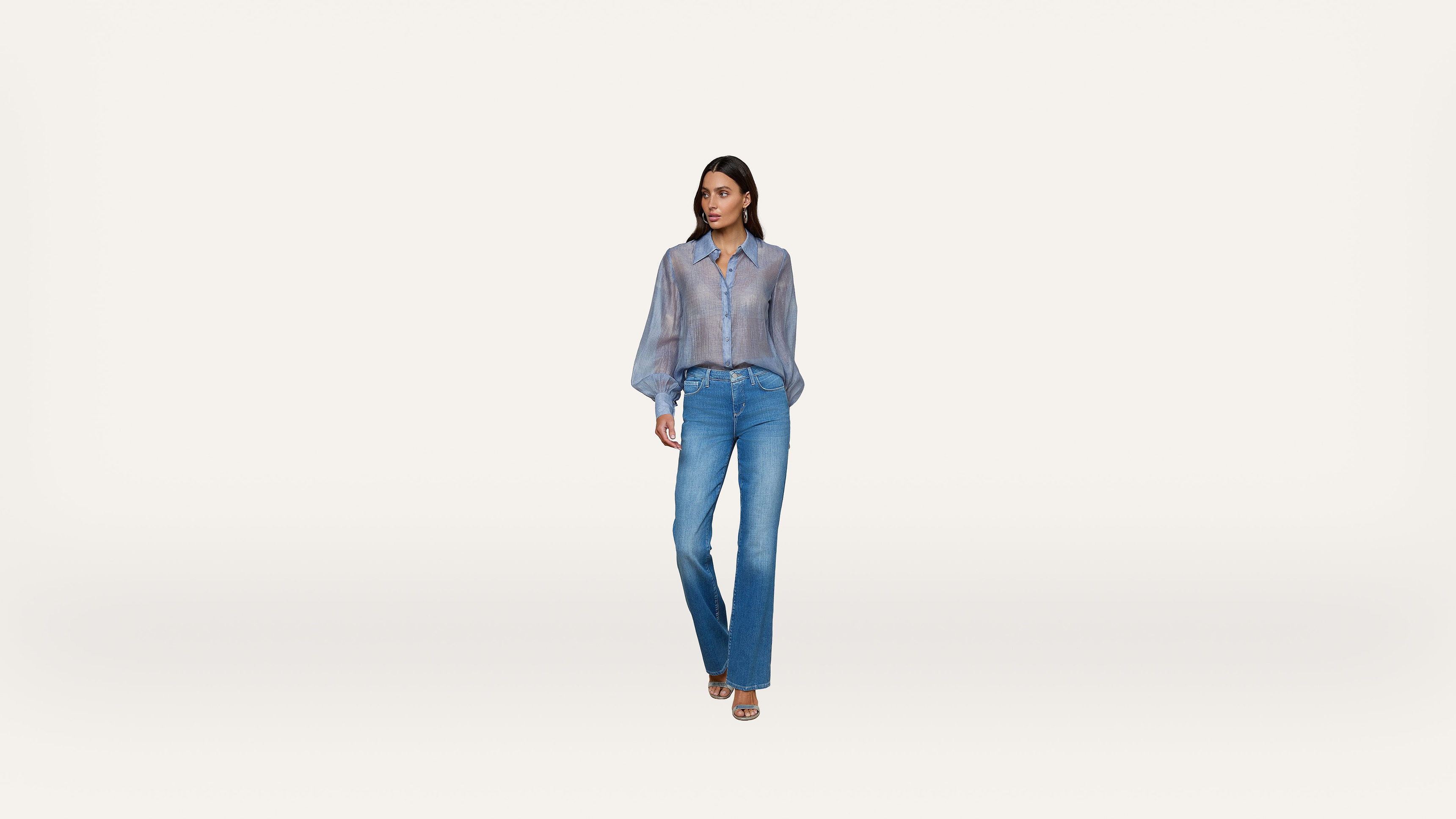 A woman stands against a plain white background, wearing a sheer blue long-sleeve shirt, blue jeans, and open-toed heels.