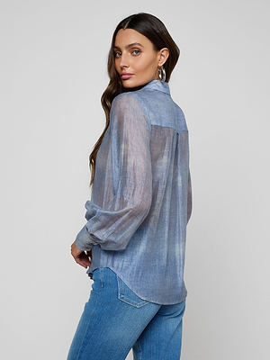 A woman with long brown hair models the Jayleen Blouse—a sheer chiffon button-down with blouson sleeves—paired with blue jeans, standing sideways and looking over her shoulder at the camera against a light background.