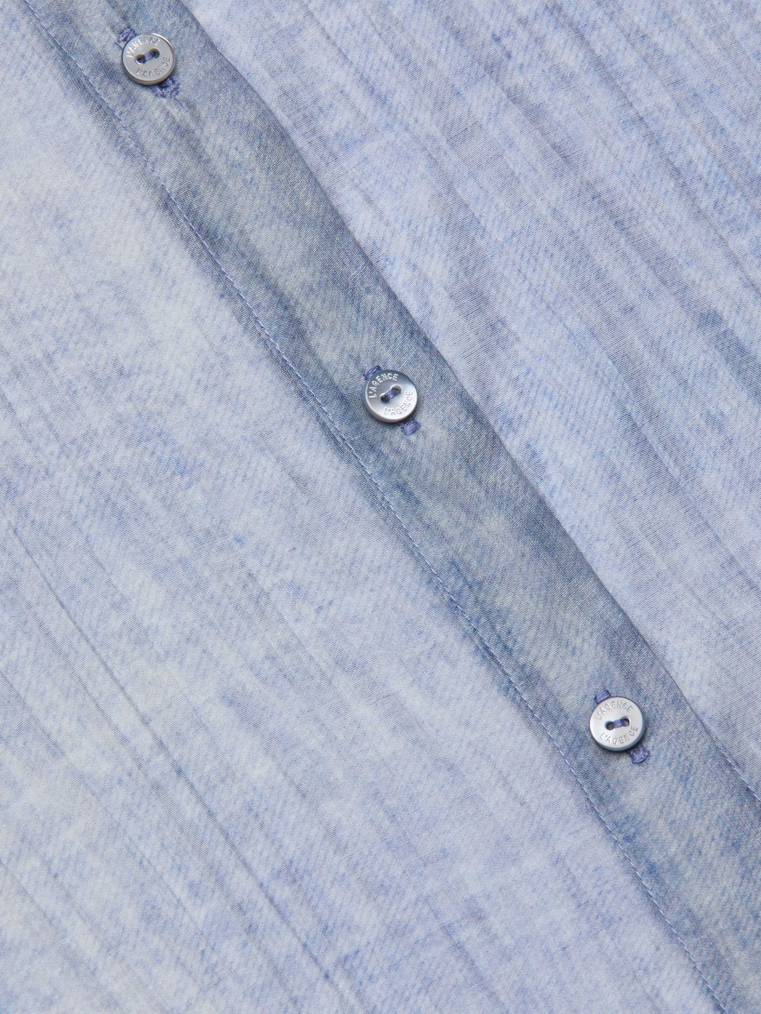 Close-up of the Jayleen Blouse: a light blue, textured button-down shirt featuring a subtle, irregular pattern and a row of white buttons with visible stitching along the vertical seam.
