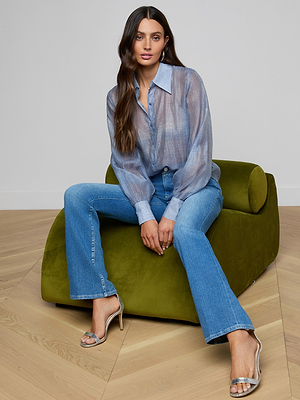 A woman with long brown hair sits on a modern olive-green chair, wearing the Jayleen Blouse—a sheer blue chiffon top with blouson sleeves—paired with blue jeans and silver high-heeled sandals, looking at the camera with a relaxed expression.