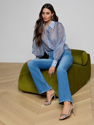 A woman with long dark hair sits on a green modern chair wearing the Jayleen Blouse, blue jeans, silver hoop earrings, and silver heeled sandals. The background has light wood flooring and a plain white wall.