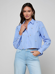 A woman with long dark hair wears the Cosette Sequined Shirt and light wash jeans, posing with one hand touching her collar against a plain light background.
