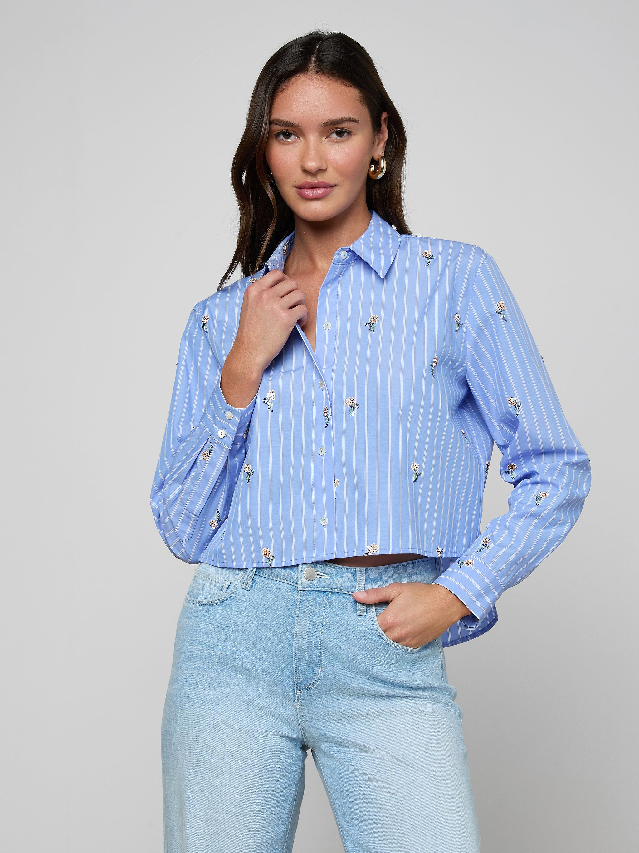 A woman with long dark hair wears the Cosette Sequined Shirt and light wash jeans, posing with one hand touching her collar against a plain light background.