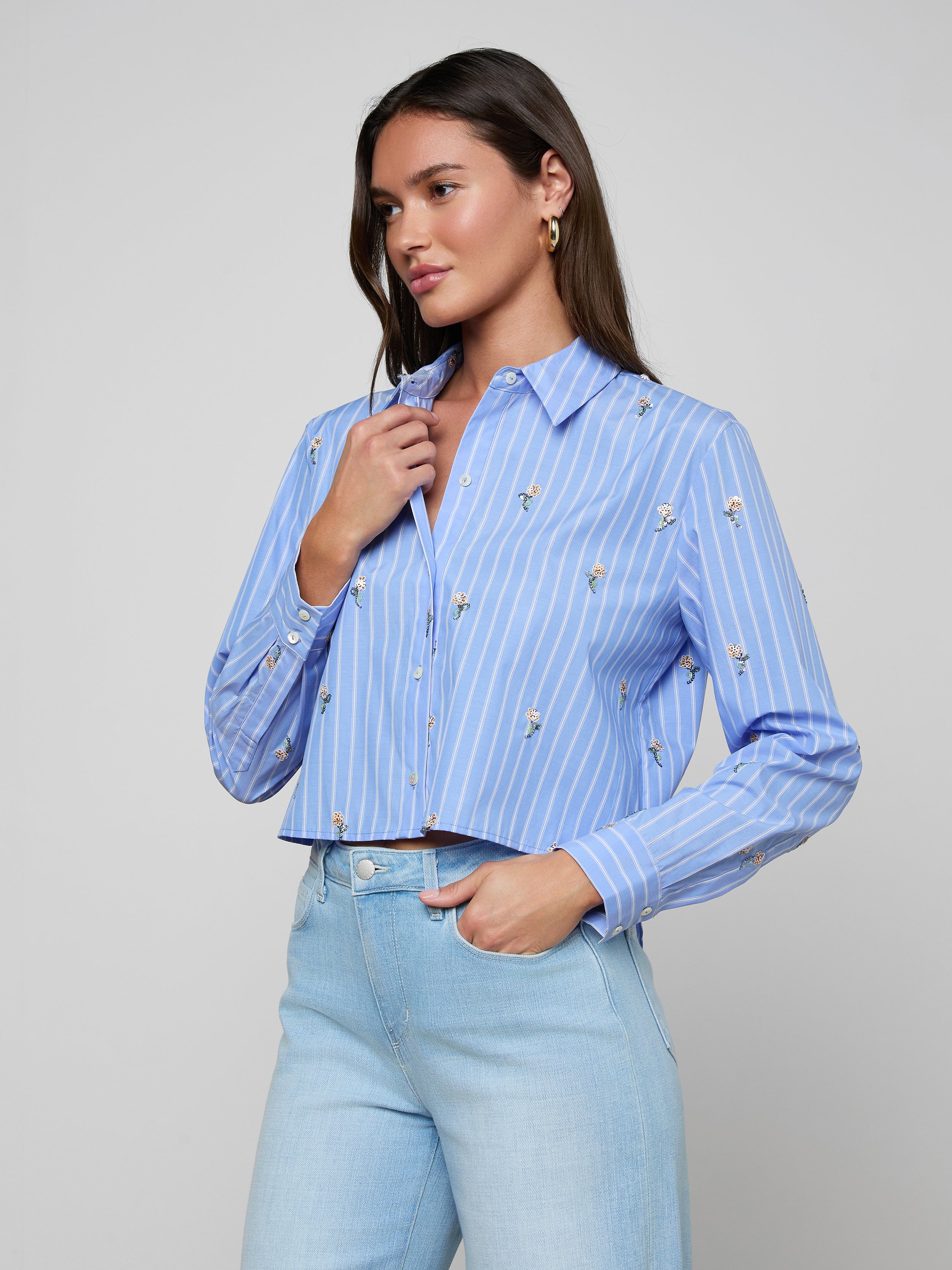 A woman wears the Cosette Sequined Shirt in blue with long sleeves, featuring white stripes and small floral patterns, and stands with one hand holding the shirt collar. She pairs it with high-waisted light blue jeans.