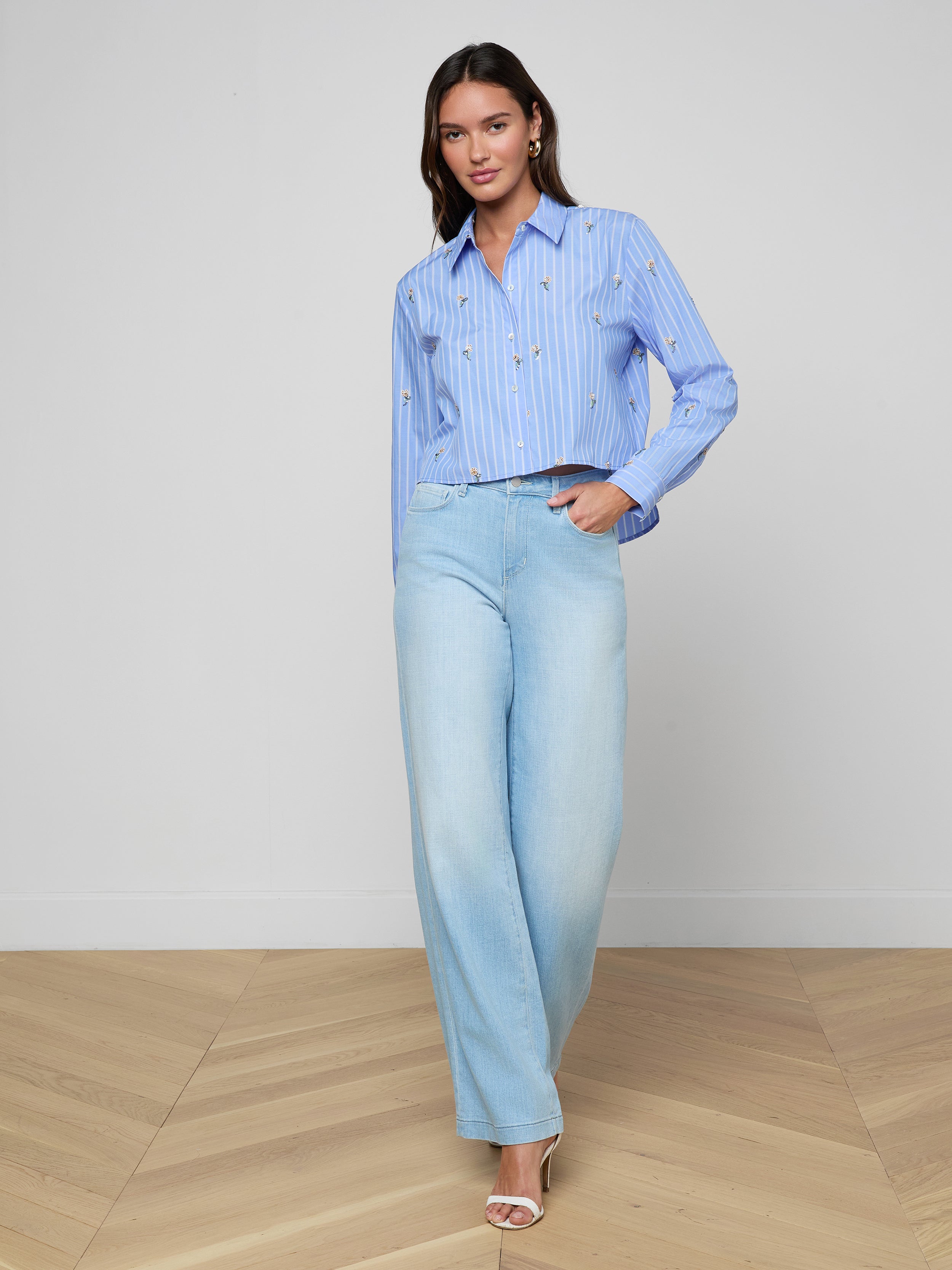 Woman standing against a plain white background, wearing the Cosette Sequined Shirt with a light blue cropped high-low hem, light blue wide-leg jeans, and white heeled sandals, with one hand in her pocket.