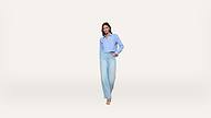 A woman stands against a plain white background, wearing a light blue button-down shirt with a pattern and light blue wide-leg jeans, paired with open-toe heels.