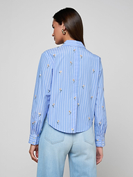 A woman is seen from behind wearing the Cosette Sequined Shirt—a blue long-sleeve poplin shirt with white vertical stripes and subtle floral prints, featuring a cropped high-low hem—paired with light blue jeans against a plain light background.