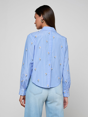 A woman stands facing away, wearing the Cosette Sequined Shirt with white vertical stripes and small embroidered designs, paired with light blue jeans. Her straight brown hair is worn down.