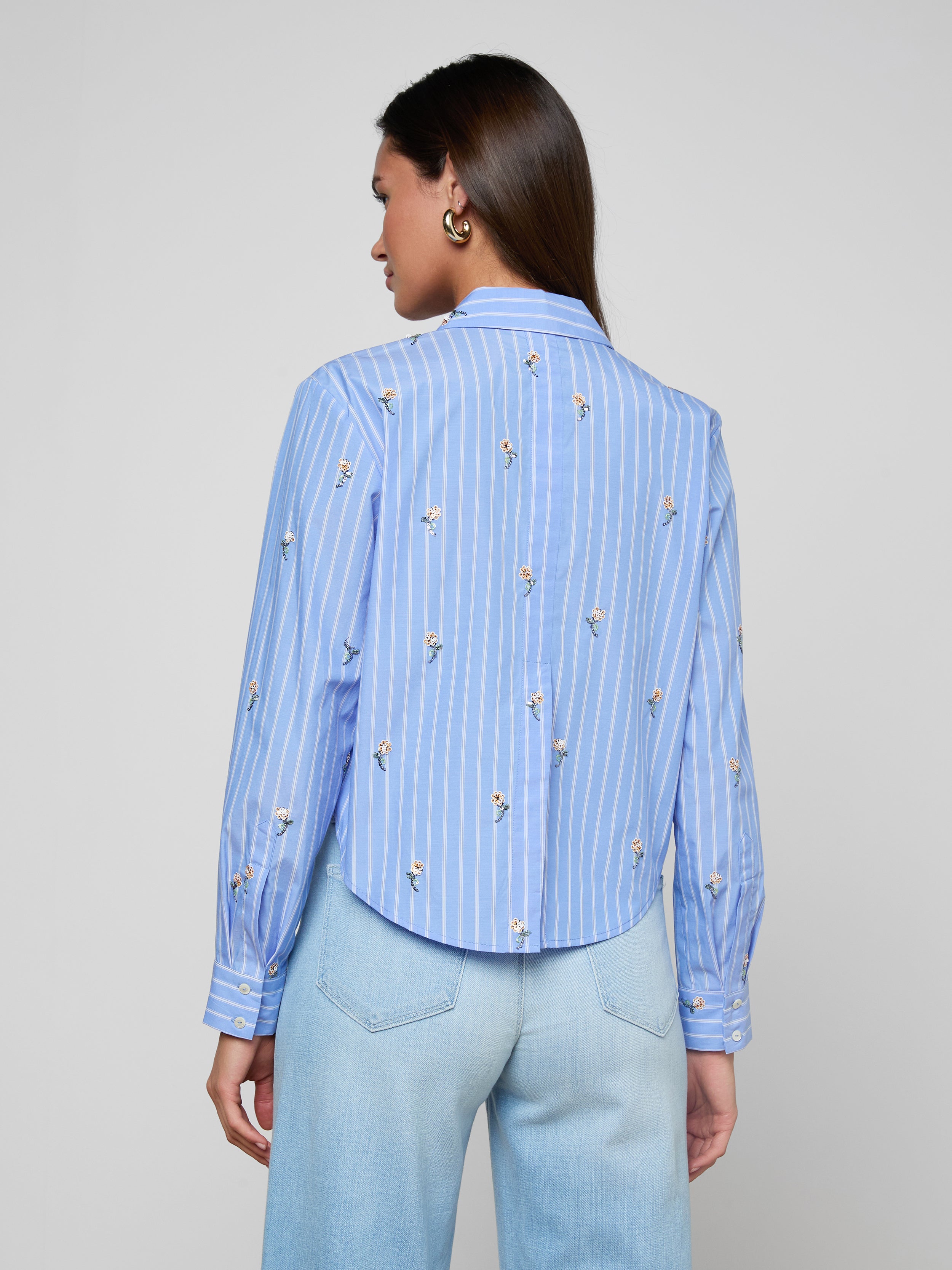 A woman is seen from behind wearing the Cosette Sequined Shirt—a blue long-sleeve poplin shirt with white vertical stripes and subtle floral prints, featuring a cropped high-low hem—paired with light blue jeans against a plain light background.