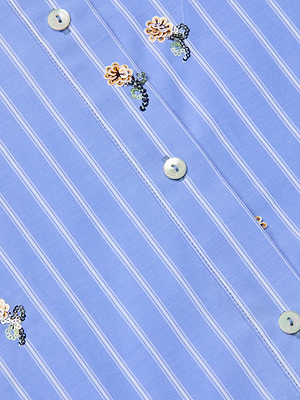 Close-up of the Cosette Sequined Shirt, a light blue striped poplin design featuring white vertical stripes, iridescent buttons, embroidered floral motifs, and delicate sequin flower embellishments scattered throughout.