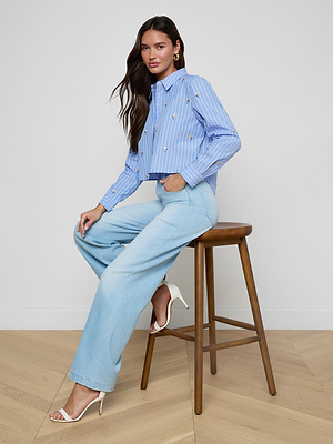 A woman with long brown hair sits sideways on a wooden stool, wearing the Cosette Sequined Shirt, light blue wide-leg jeans, and white high-heeled sandals against a plain background.