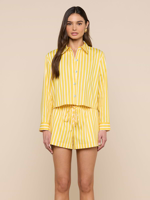 A woman stands facing forward, wearing the Cosette Shirt—a matching yellow and white vertical striped organic cotton button-up—paired with drawstring shorts against a plain light background.