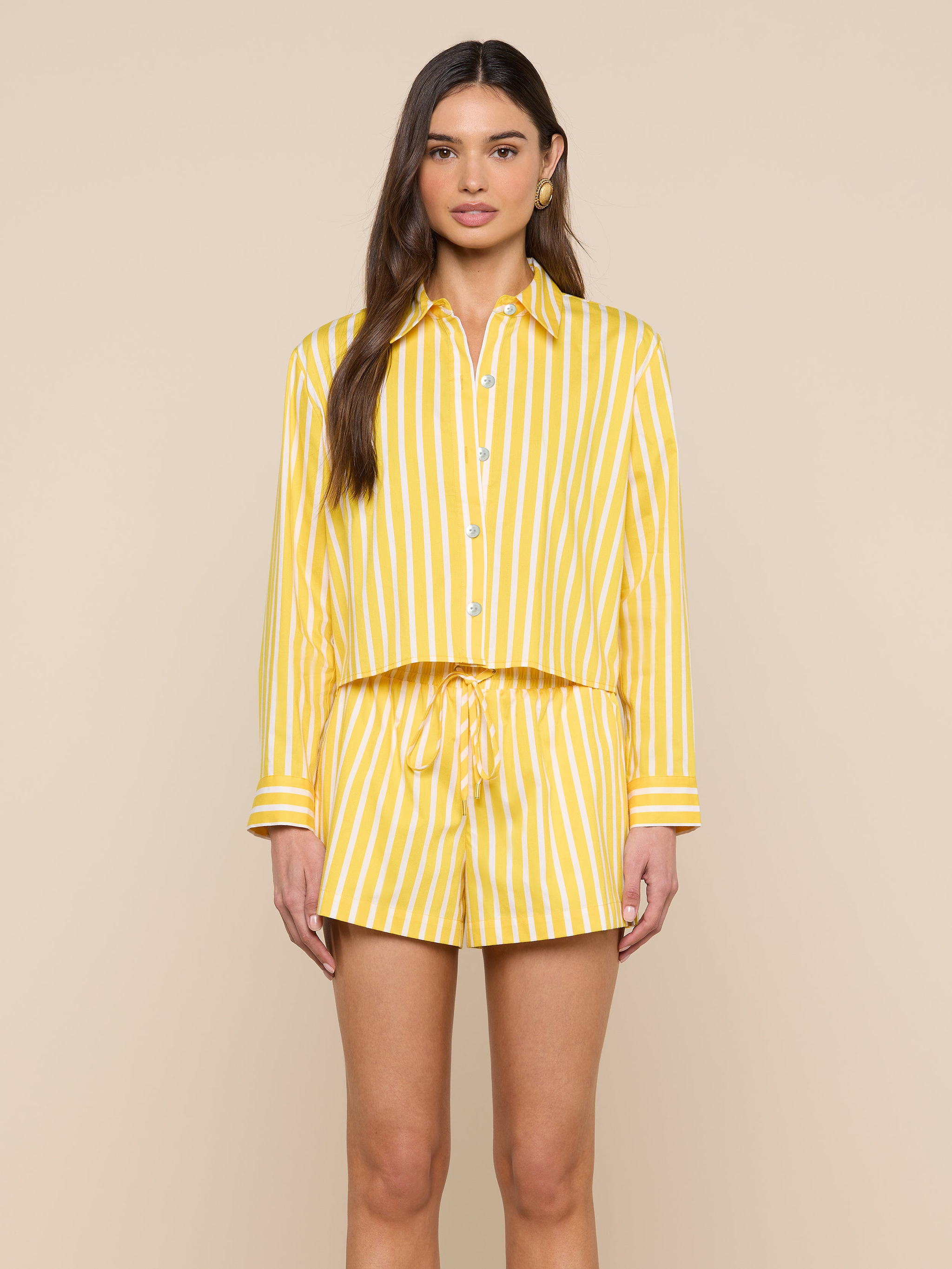 A woman stands facing forward, wearing the Cosette Shirt—a matching yellow and white vertical striped organic cotton button-up—paired with drawstring shorts against a plain light background.