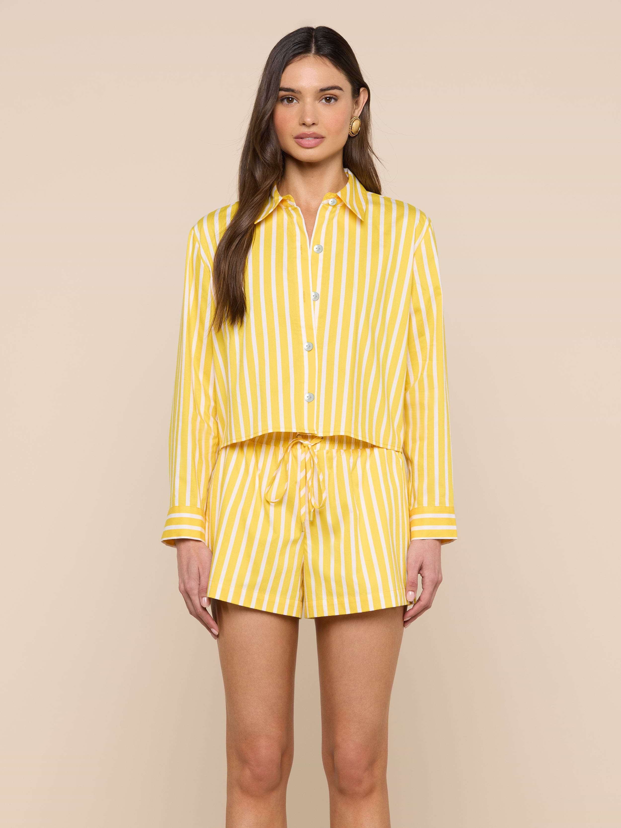 A woman stands facing forward, wearing the Cosette Shirt—a matching yellow and white vertical striped organic cotton button-up—paired with drawstring shorts against a plain light background.