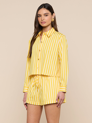 A woman stands against a beige background wearing the Cosette Shirt—a classic, yellow and white vertically striped button-up—paired with matching drawstring shorts. She has long brown hair and accessorizes with gold hoop earrings.