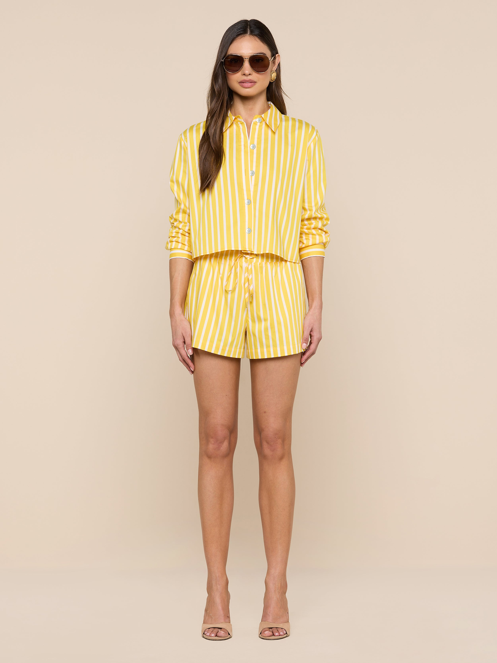 A woman wearing sunglasses and the Cosette Shirt, a yellow and white vertical striped tailored shirt with sleeves rolled up, paired with matching shorts and nude high-heeled sandals, stands against a plain white background.