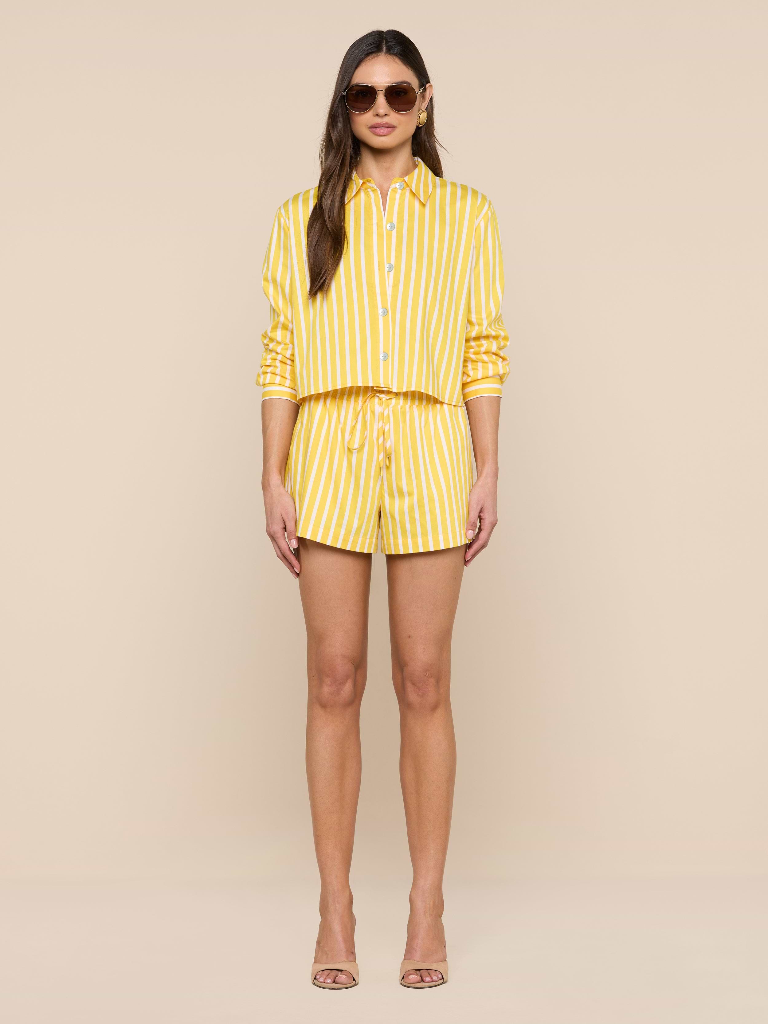 A woman wearing sunglasses and the Cosette Shirt, a yellow and white vertical striped tailored shirt with sleeves rolled up, paired with matching shorts and nude high-heeled sandals, stands against a plain white background.