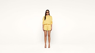 Woman stands against a plain white background wearing a yellow striped shirt, matching shorts, heeled sandals, and sunglasses. Her arms rest at her sides and she faces forward, looking at the camera.