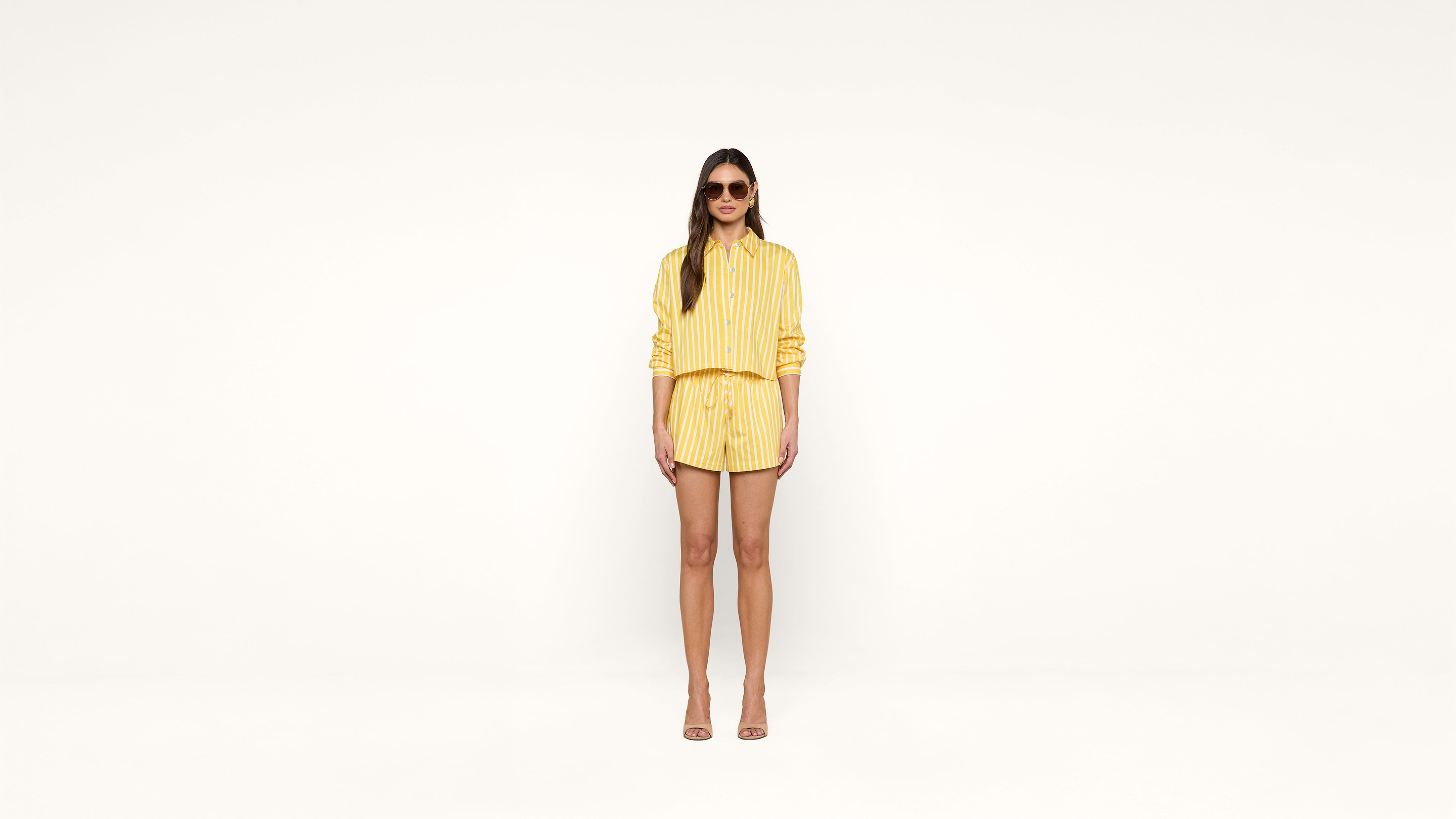 Woman stands against a plain white background wearing a yellow striped shirt, matching shorts, heeled sandals, and sunglasses. Her arms rest at her sides and she faces forward, looking at the camera.