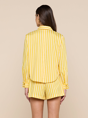 A woman stands facing away, wearing the Cosette Shirt—an organic cotton-blend poplin top with yellow and white vertical stripes, a cropped high-low hem, and matching shorts against a light beige background.