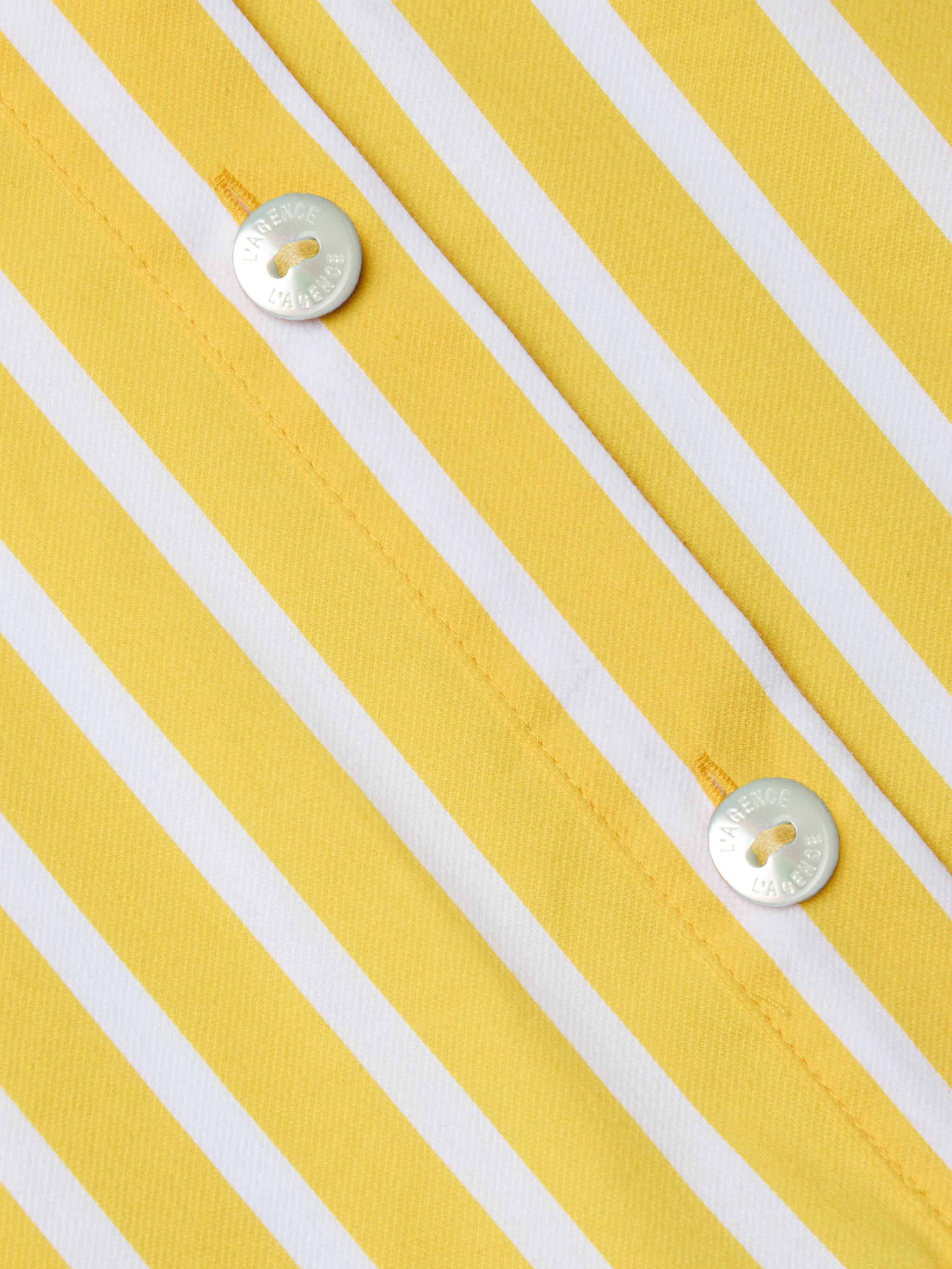 Close-up of the Cosette Shirt, a tailored piece made from organic cotton in yellow and white vertical stripes, featuring two shiny silver buttons engraved with text and sewn on with yellow thread.