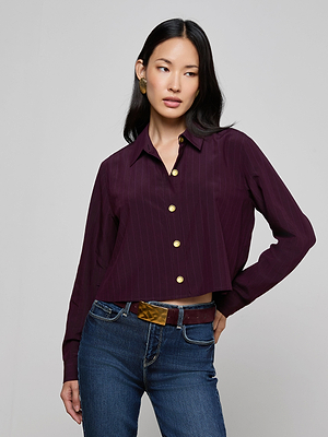A woman with long dark hair models the Cosette Pinstriped Shirt in burgundy, paired with blue jeans, standing against a light gray background and looking to her left with one hand on her hip.