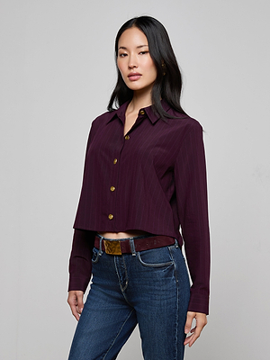 A woman with long dark hair models the Cosette Pinstriped Shirt, paired with blue jeans and a belt. She stands facing slightly to the side against a plain light gray background.
