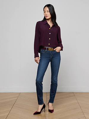 A woman with long dark hair poses against a plain light gray wall, wearing the Cosette Pinstriped Shirt, blue jeans, a brown belt, and burgundy high heels. One hand is in her pocket.
