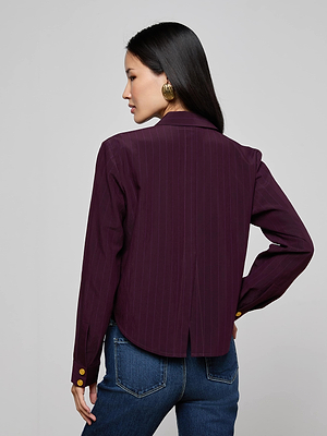 A woman with long black hair wears the Cosette Pinstriped Shirt with gold buttoned cuffs and blue jeans, standing facing away and glancing to the side against a plain background.