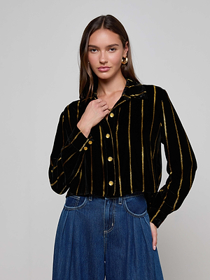 A woman with long brown hair wears the Cosette Velvet Shirt—black velvet with metallic gold stripes and gold buttons—paired with high-waisted blue jeans. She stands against a plain light gray background, looking at the camera.