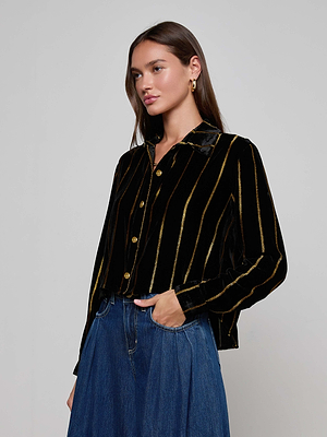 A woman with long brown hair wears the L'AGENCE Cosette Velvet Shirt with gold vertical stripes and wide-leg blue jeans, standing against a light gray background. The shirt’s oversized fit adds a modern edge to her look.