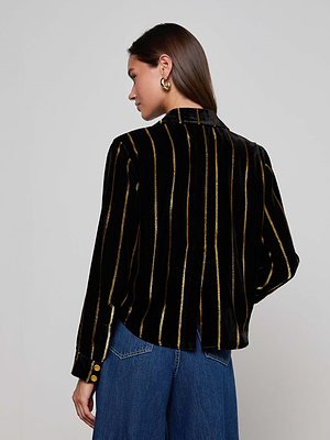A woman with straight brown hair models the L'AGENCE Cosette Velvet Shirt—an oversized black velvet top with metallic gold stripes—paired with blue jeans, standing with her back to the camera against a plain light background.