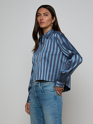 A woman with long brown hair wears the Cosette Satin Shirt in blue stripes with a high-low hem and light blue jeans, standing against a plain light gray background.