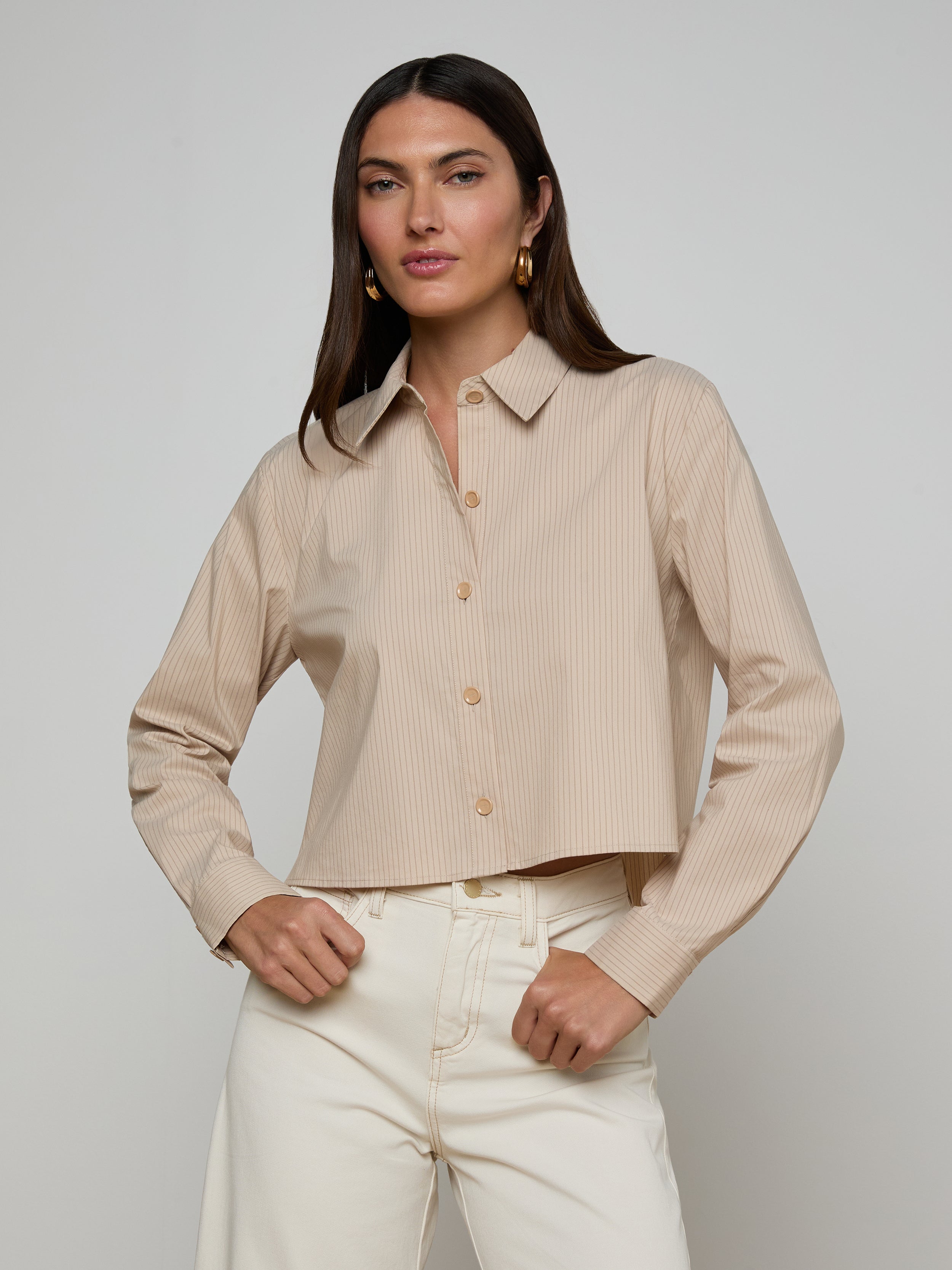 A woman stands with her hands on her hips, wearing the Cosette Shirt—a tailored, beige long-sleeve button-up—paired with off-white pants. She has long, straight brown hair and faces forward against a plain light background.