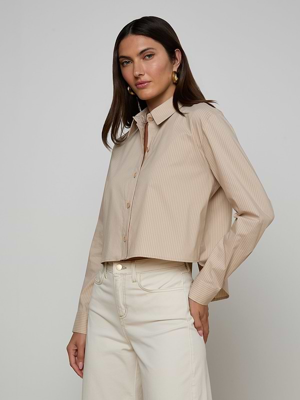 Wearing the Cosette Shirt, a beige, long-sleeve organic cotton top with subtle stripes, a woman pairs it with high-waisted cream pants. She stands against a plain light background, looking confidently at the camera.