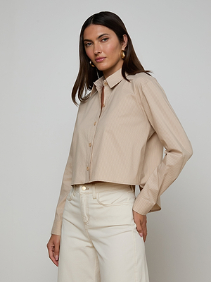 A woman with long brown hair wears the tailored, tan Cosette Shirt made from organic cotton, paired with high-waisted cream jeans. She stands against a plain light gray background, looking confidently at the camera.