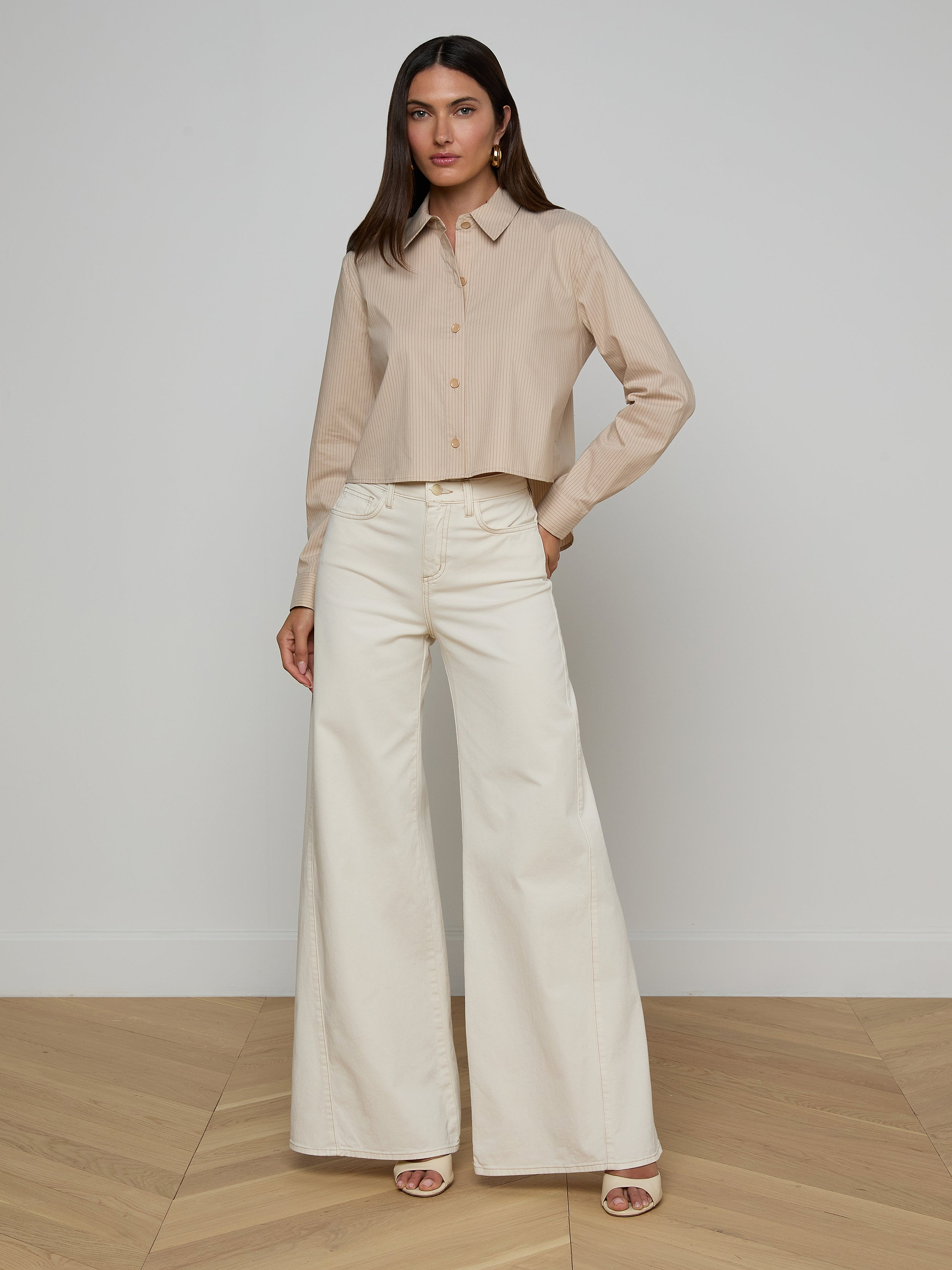 A woman stands against a plain white background, wearing the Cosette Shirt made from organic cotton with long sleeves, paired with high-waisted, wide-leg white pants. She has long dark hair and is wearing open-toed heels.