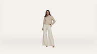 A woman stands against a plain white background, wearing a beige button-up shirt and high-waisted, wide-leg white pants with beige platform heels. She has one hand in her pocket and a neutral expression.