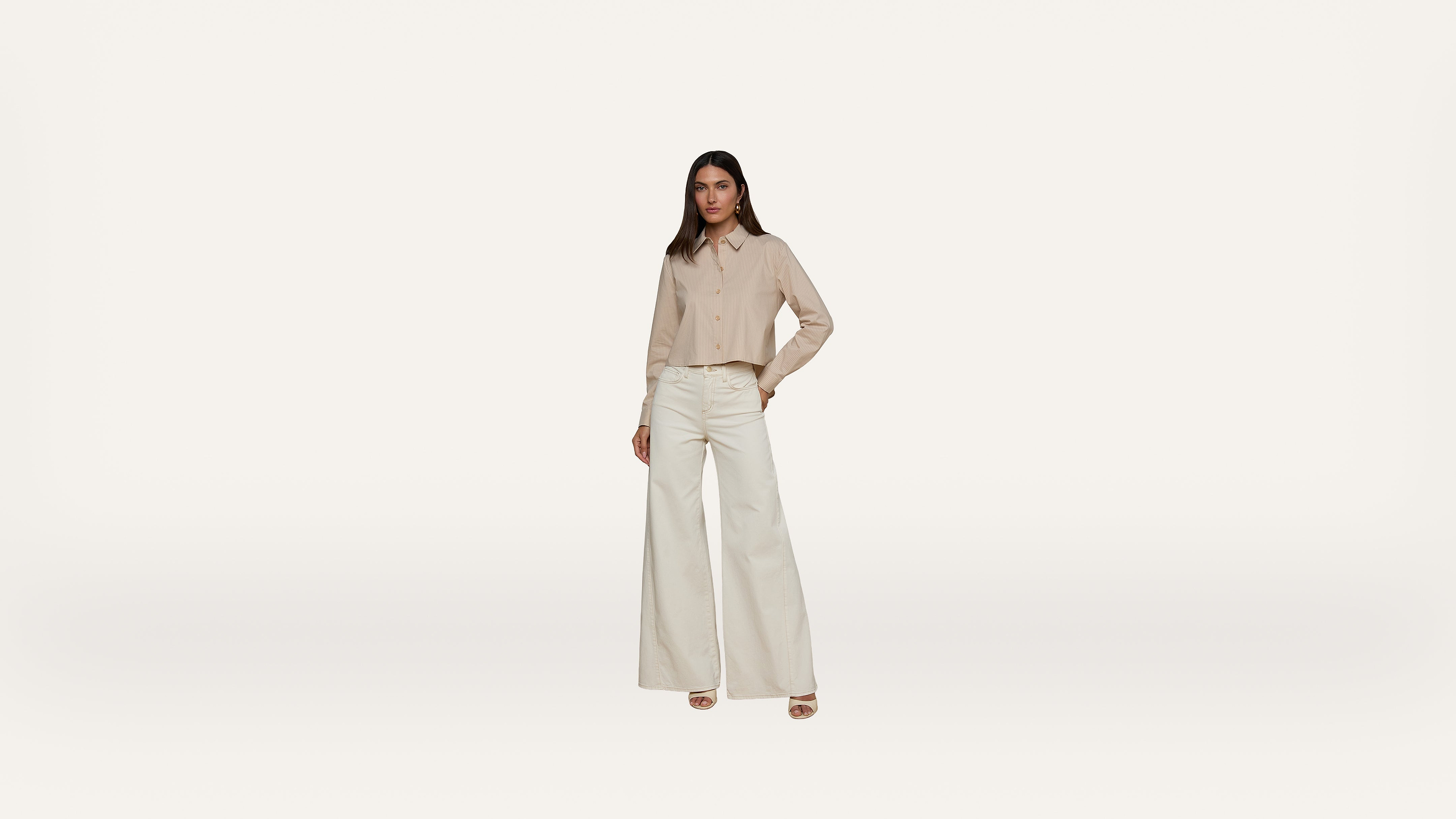 A woman stands against a plain white background, wearing a beige button-up shirt and high-waisted, wide-leg white pants with beige platform heels. She has one hand in her pocket and a neutral expression.