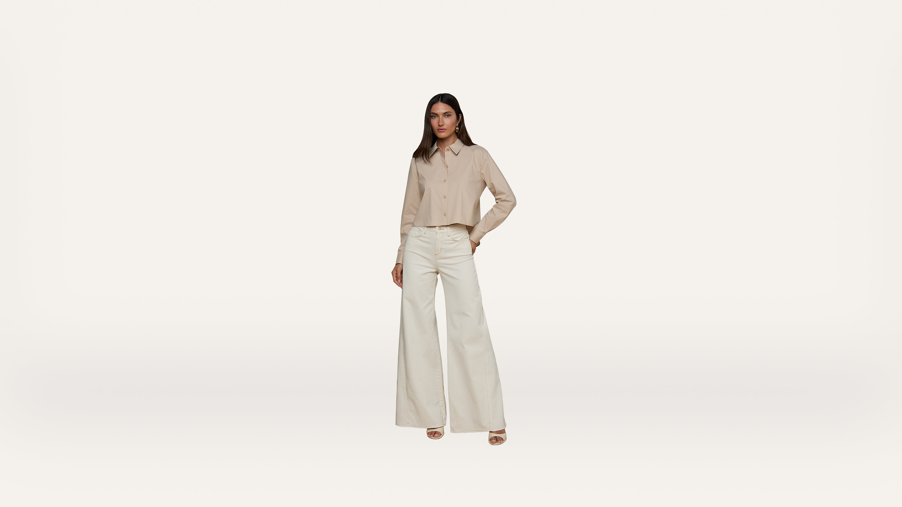 A woman stands against a plain white background, wearing a beige button-up shirt and high-waisted, wide-leg white pants with beige platform heels. She has one hand in her pocket and a neutral expression.