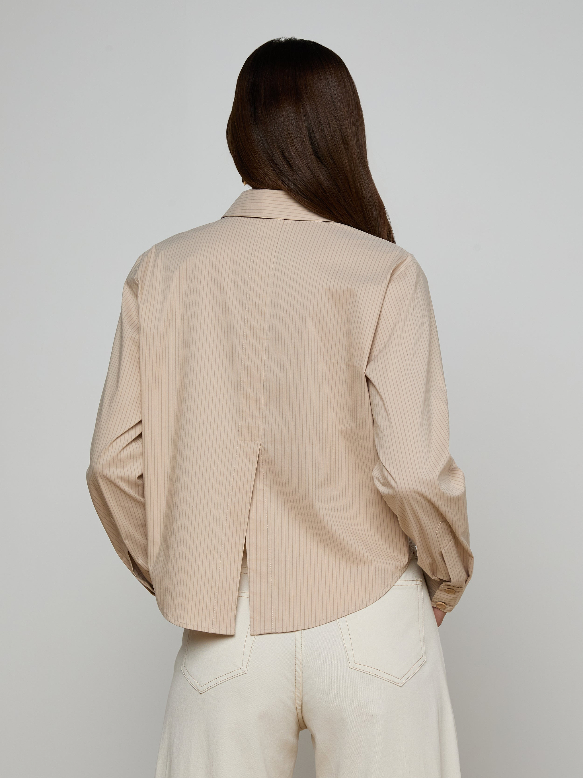 A person with long brown hair, seen from the back, wears the Cosette Shirt—a beige, collared, tailored shirt with long sleeves and a back slit—paired with white pants. The background is plain and light-colored.
