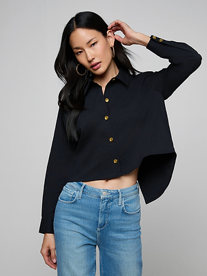 A woman with long black hair confidently poses in front of a plain background wearing the Cosette Shirt, a tailored black button-up with gold buttons, paired with light blue jeans. One hand touches her head.
