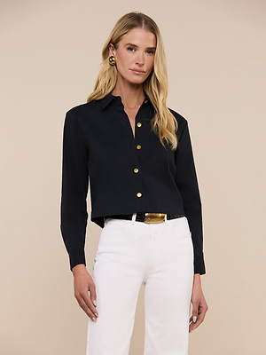 A woman with long blonde hair wears the Cosette Shirt—black, tailored, with gold buttons—paired with white pants, a black belt featuring a gold buckle, and gold earrings. She stands against a plain beige background.