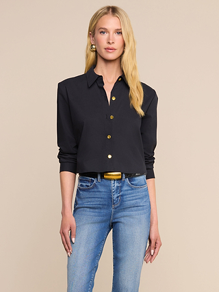 A woman with long blonde hair wears the Cosette Shirt—a classic, tailored black button-up crafted from an organic cotton-blend—paired with blue jeans, a black belt featuring a gold buckle, and gold earrings, standing against a beige background.