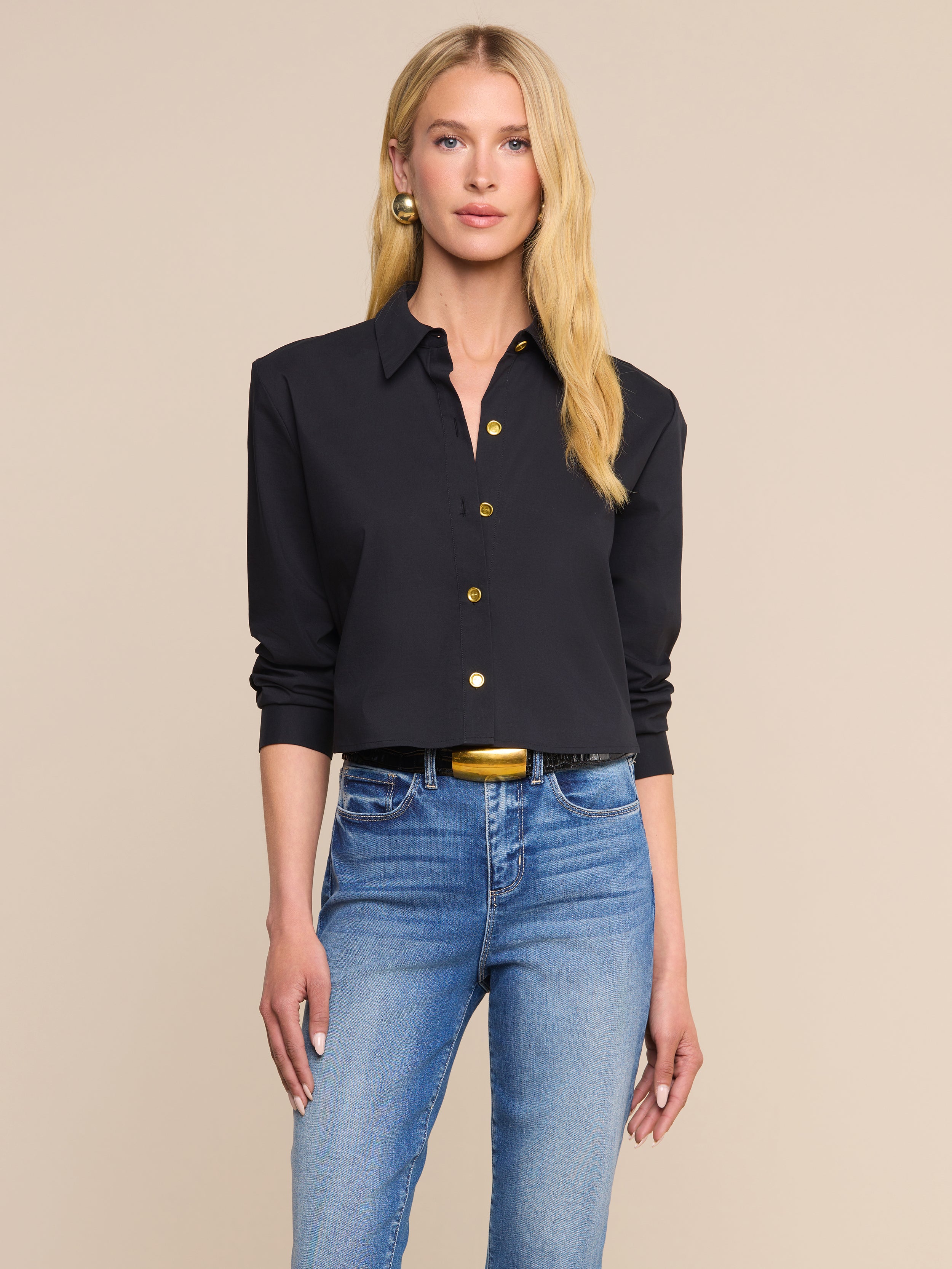 긴 금발 머리를 한 여성이 유기농 면 혼방 소재로 제작된 클래식하고 정교한 핏의 검은색 단추 셔츠인 ‘코제트 셔츠’를 입고, 청바지와 금색 버클이 달린 검은색 벨트, 금색 귀걸이를 매치한 채 베이지색 배경 앞에 서 있다.